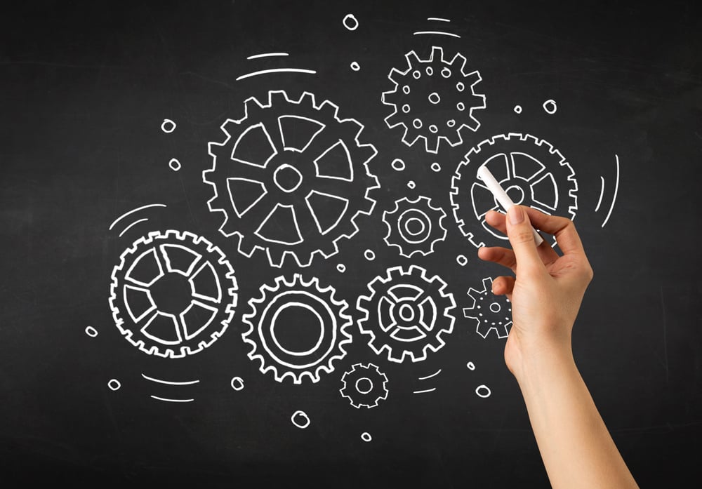 Female hand holding white chalk in front of a blackboard with gears drawn on it